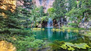 The vibrant turquoise waters of Hanging Lake ripple gently as waterfalls cascade over moss-covered cliffs, surrounded by lush greenery.