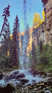 View from behind the waterfall at Hanging Lake, where bright yellow and orange trees are bathed in the soft glow of morning sunlight.