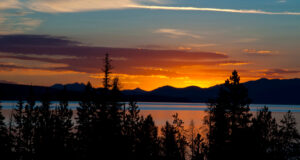 A vibrant orange sunrise reflects over calm water with mountains silhouetted in the background, casting a peaceful glow on the scene.